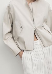 Person iført en lys beige zip-up jakke med tryklåslommer, parret med hvide højtaljede bukser med fine lodrette striber. Hænderne i lommerne.