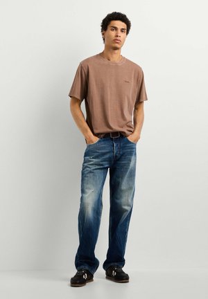Giovane uomo in piedi con le mani nelle tasche, indossa una maglietta marrone, jeans blu sbiaditi e sneakers nere su uno sfondo chiaro e uniforme.