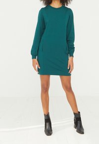 Femme portant une robe mini à manches longues turquoise avec deux poches latérales, assortie à des bottines noires, se tenant sur un sol blanc devant un fond blanc.