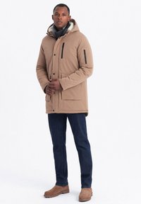 Parka beige isolato con cappuccio, tasche laterali, dettagli in zip neri, abbinata a jeans blu scuro e stivali in suede marroni.