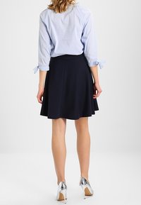 Marineblauer ausgestellter Rock kombiniert mit einer hellblau gestreiften Bluse mit gebundenen Ärmeln, ergänzt durch glänzende silberne High Heels.