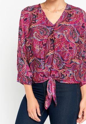 Femme portant un chemisier rose à motif paisley avec un col en V et un lien à l'avant, associé à un jean bleu foncé.