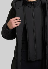 Chaqueta acolchada negra con un alto cuello, que cuenta con múltiples cremalleras frontales y una capa interior desmontable, textura suave y un corte ajustado.