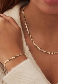 Goldfarbene Metallkette und -armband mit einem strukturierten, runden Glieddesign, getragen über einem weichen, cremefarbenen Pullover.