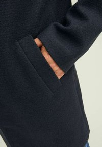 Cappotto di lana scuro con una tasca laterale e una mano parzialmente inserita. Il materiale sembra morbido e testurizzato. Design semplice e ben tagliato.