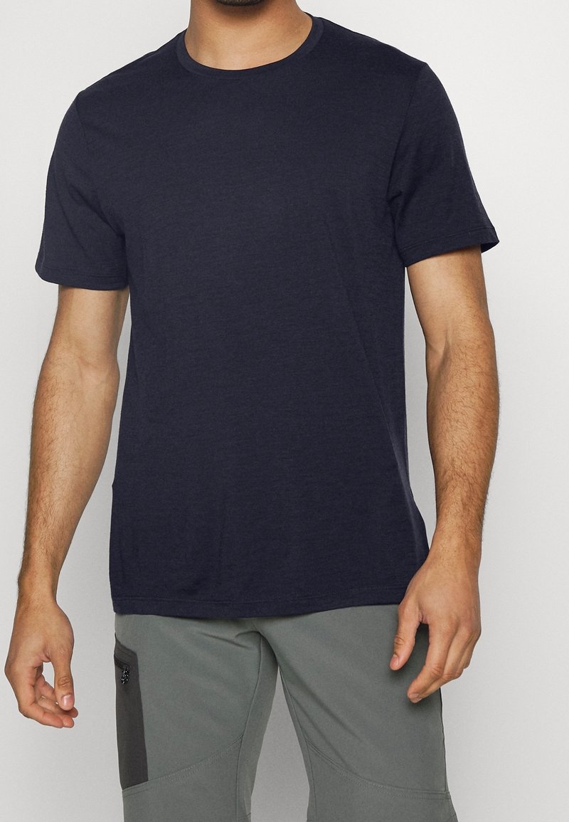 Mand iført en ensfarvet mørk marineblå kortærmet t-shirt med rund hals og lysegrå bukser med en sort lynlåslomme.