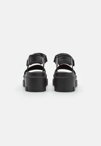 Paire de sandales plateformes noires épaisses avec des brides à la cheville et des talons ouverts, montrées de dos sur fond blanc.