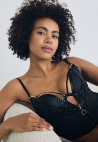Sort læder bralette med et struktureret design, der har dekorative guld accenter og krydsede stropper. Glat tekstur, tætsiddende form.