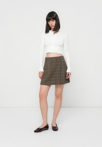 Woman with straight black hair wearing a white cropped zip hoodie, brown plaid skirt, and dark red ballet flats, standing against a white background.