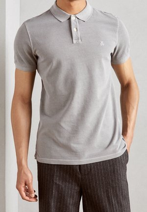 Man wearing light grey short-sleeve polo shirt with two buttons and dark pinstriped pants, standing with one hand in pocket.