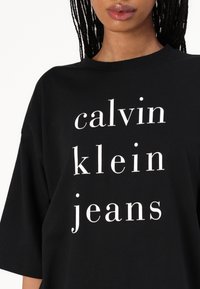 Černé tričko s krátkými rukávy, na kterém se nachází výrazný bílý text: "calvin klein jeans" uspořádaný do sloupcového designu, vyrobené z bavlny s hladkým povrchem.