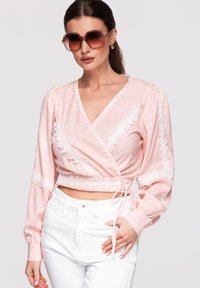 Femme portant de grandes lunettes de soleil marron, un chemisier cache-cœur rose orné de dentelle et un pantalon blanc taille haute, posant avec une main sur la hanche.