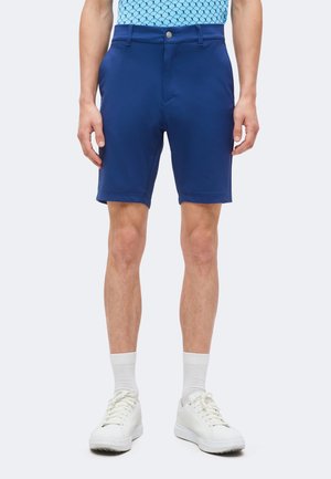 Personne portant un short bleu vif, une chemise bleu clair à motifs, des chaussettes blanches et des baskets blanches, debout devant un fond clair et uni.