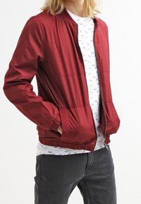 Giacca bomber rossa, leggera, con zip frontale, polsini elasticizzati e orlo a coste. Indossata sopra una camicia bianca con motivi neri e pantaloni grigi.