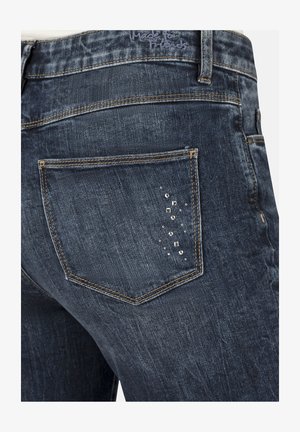 Dark blue denim jeans featuring a back pocket with silver embellishments, distressed texture, and contrast stitching along the edges.