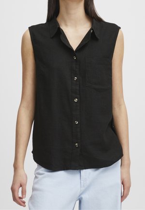 Vrouw draagt een zwarte mouwloze blouse met knopen en een voorzak, en lichtblauwe spijkerbroek, staand tegen een effen achtergrond.