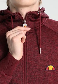 Maroon zip-up hoodie made of textured fabric, featuring a logo on the left chest and adjustable drawstrings on the hood. Silver zipper detail.