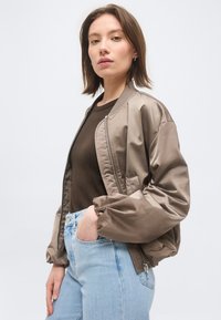 Jauna sieviete, kas valkā spīdīgu bēša krāsas bomberjaku, tumši brūnu kreklu un gaiši zilas džinsas, pozējot ar vienu roku kabatā pret vienkāršu fonu.