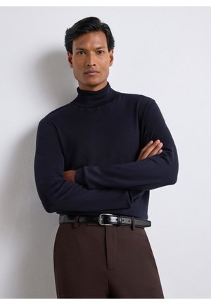 Uomo con le braccia incrociate che indossa un maglione nero a collo alto e pantaloni marroni con una cintura nera davanti a una parete bianca liscia.