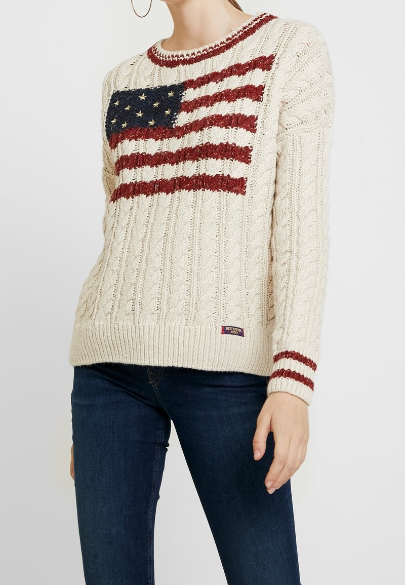 Femme portant un pull crème à torsades avec un motif du drapeau américain et des rayures bordeaux sur le col et les manches, associé à un jean foncé.