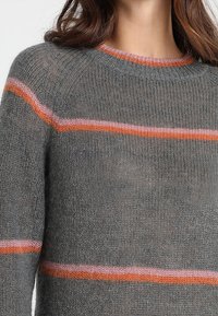 Grå stickad tröja med rund halsringning, prydd med horisontella ränder i rosa och orange. Mjuk textur, lätt och något genomskinlig.