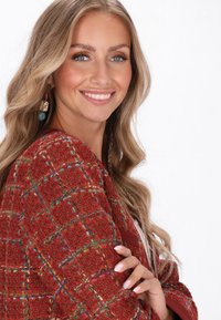 Giacca in tweed rosso con scacchi multicolori, caratterizzata da un tessuto testurizzato. Il modello indossa orecchini e trucco leggero con capelli lunghi e ondulati.