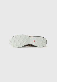 Semelle de chaussure de sport noire et marron avec une semelle extérieure en caoutchouc blanc texturé, dotée de crampons multidirectionnels et d'un détail en accent rouge.