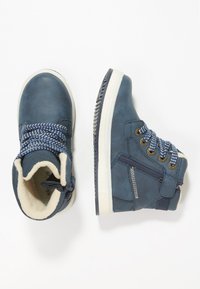 Botas de tobillo de cuero azul con forro de piel blanca, cordones con patrón azul y blanco, ojales dorados y suela blanca. Incluye detalle de cierre lateral.