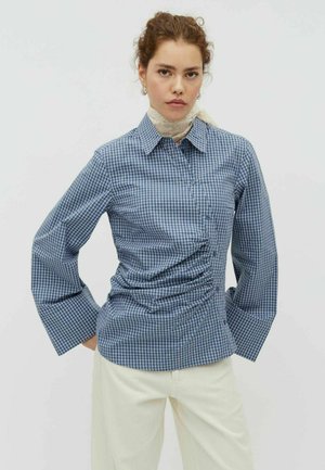 Frau mit lockigem Haar, die ein blau kariertes, gerafftes Hemd, weiße Hose, ein Spitzenhalsband und Creolen vor einem einfarbigen Hintergrund trägt.