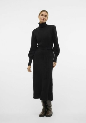 Schwarzes Rippstrickkleid mit hohem Kragen, tailliertem Schnitt, langen Puffärmeln und Midi-Länge. Kombiniert mit kniehohen Stiefeln.