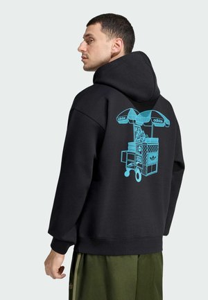 Mężczyzna w czarnej bluzie z kapturem z niebieską grafiką wózka na buty Adidas na plecach, w połączeniu z zielonymi spodniami na jednolitym jasnym tle.