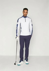 Balta ilgomis rankovėmis golfinė marškinė su mėlynais akcentais, derinami su mėlynomis kelnėmis ir baltomis golfinėmis bateliais. Modelis laiko golfą.