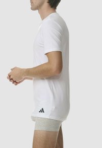 T-shirt bianco a maniche corte in cotone, con un logo discreto a tre strisce nere sul lato sinistro in basso, indossato con pantaloni boxeur grigi.