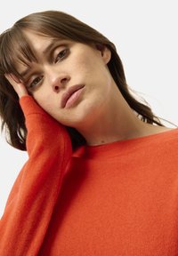 Pull rouge en tricot doux avec une coupe décontractée, un col légèrement large et des manches longues. La texture semble lisse et uniforme.