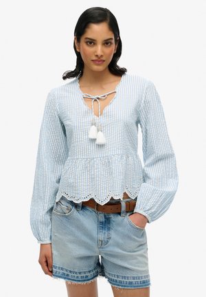 Blusa de manga larga a rayas azul claro con un dobladillo en ondas, cuello atado con borlas blancas, combinada con pantalones cortos de mezclilla.