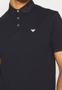 Svart piké t-shirt i bomull, med en knäppt krage med "EMP"-text, en liten vit logotyp på vänster bröst och korta ärmar.