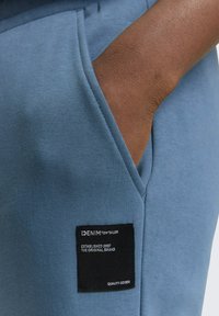 Niebieska bawełniana bluza z boczną kieszenią i czarną metką z białym nadrukiem. Materiał ma gładką fakturę i delikatną rozciągliwość.