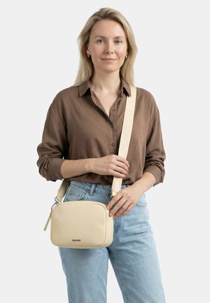 Blond naine, kes kannab pruunikat särki ja helehalli teksapükse, hoiab käes kreemivärvi ristihoidikut laiade rihmadega.