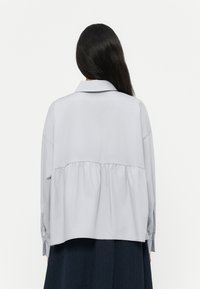 Hellgraue Bluse mit langen Ärmeln und einem Kragen. Der untere Saum ist gerafft und elastisch, was einen lässigen Sitz ermöglicht. Glatte Textur.