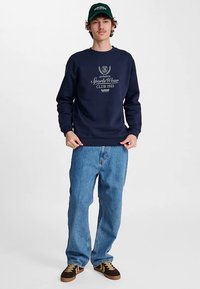 Námořnická mikina s žebrovaným výstřihem a manžetami, zdobená bílým textem a logem. Kombinována se světle modrými džíny a hnědými teniskami.