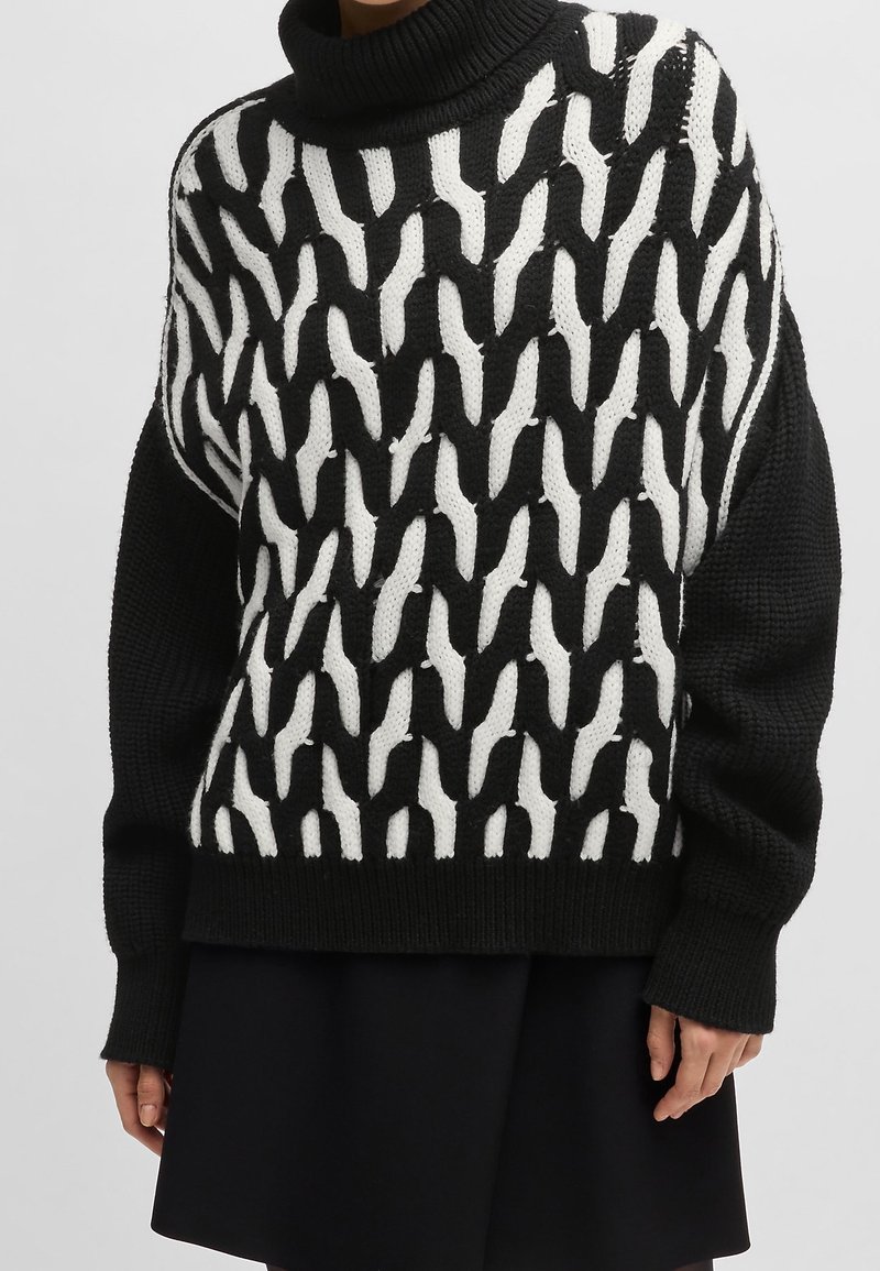 Pull en grosse maille noir et blanc avec des motifs ondulés mélangés, manches longues et col roulé épais porté avec une jupe noire.