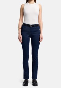 Jeans ajustados de color azul oscuro con una cintura alta, una textura suave y un diseño de cinco bolsillos, combinados con una blusa blanca sin mangas y un cinturón negro.