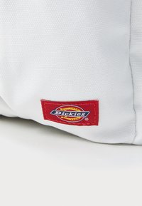 White textured fabric bag featuring a red and blue "Dickies" logo patch on the side. Compact design with no visible hardware.