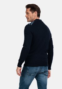 Marineblauwe gebreide trui met een gestructureerd blauw patroon op de schouders. Ribgebreide manchetten en zoom, gedragen met blauwe spijkerbroek, wat een aansluitend ontwerp toont.