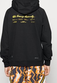 Felpa nera con testo giallo stampato sulla parte posteriore, con cappuccio, maniche lunghe e etichetta Dri-FIT sull'orlo. Abbinata a pantaloni corte a fantasia.