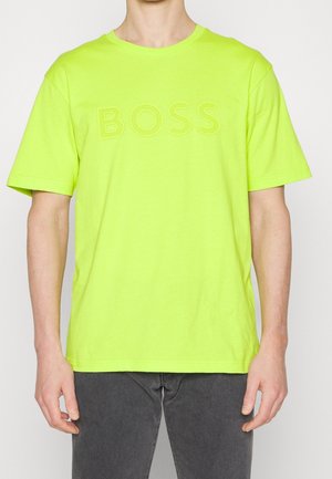 Osoba nosząca jasną neonowozieloną koszulkę z krótkim rękawem z napisem "BOSS" wytłoczonym na piersi, zestawioną z ciemnoszarymi spodniami.