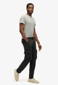 Grijze T-shirt met korte mouwen, relaxed fit, gecombineerd met zwarte cargobroek met meerdere zakken. Draagt lichte sneakers.