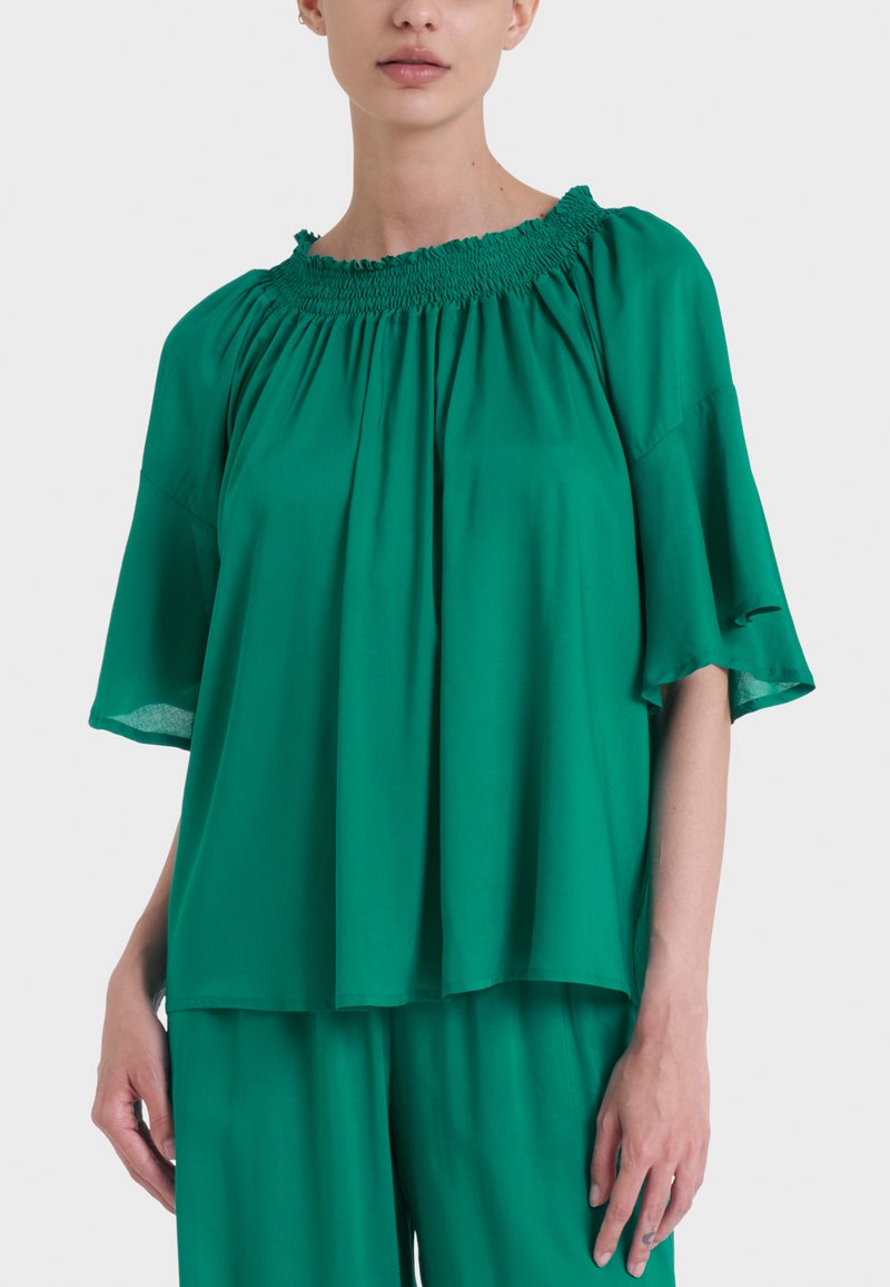 Blusa verde, ampia e morbida, realizzata in tessuto liscio. Presenta un colletto arricciato e maniche corte e larghe. Design leggero e traspirante.