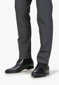 Scarpe da uomo in pelle nera con finitura liscia, dotate di punta arrotondata e lacci ridotti al minimo, abbinate a pantaloni grigi a gamba affusolata.