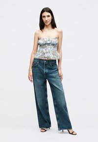 Kvinne som står iført en blomstret topp med spaghettistropper, vide blå jeans og svarte åpne hæler mot en ensfarget bakgrunn.